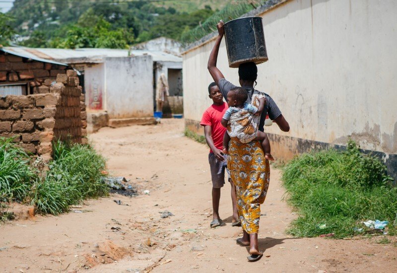 A woman in Malawi walks with a water container on her head and a baby on her back