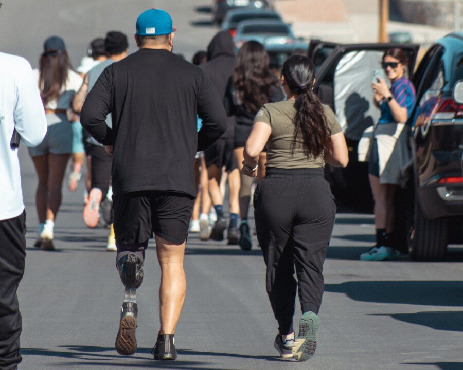A photo shot from behind a group of runners on the street. Two runners are in focus, on the left is a man with a running prosthesis on his left leg and on the right is a woman.  