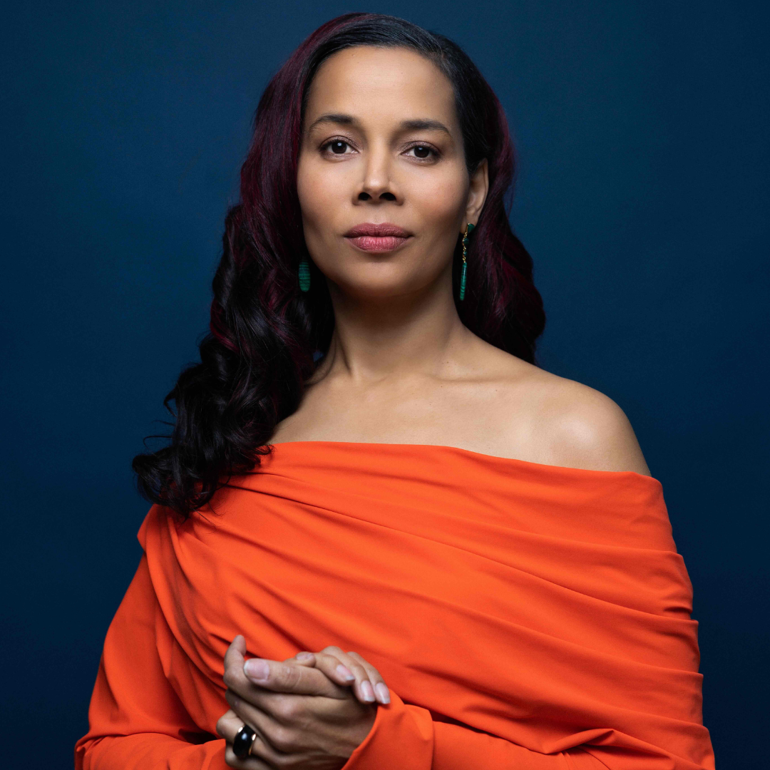 Rhiannon Giddens stands in an off the shoulder orange dress against a dark blue background with her hair down pushed over one shoulder