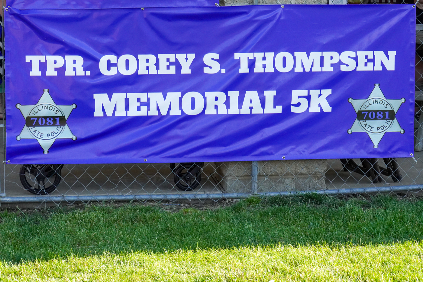 Image of a large blue banner with white text hung across a chain link fence. The text says "Tpr. Corey S. Thompsen Memorial 5k" and an silver Illinois State Police badge with #7081 is on either bottom corner.