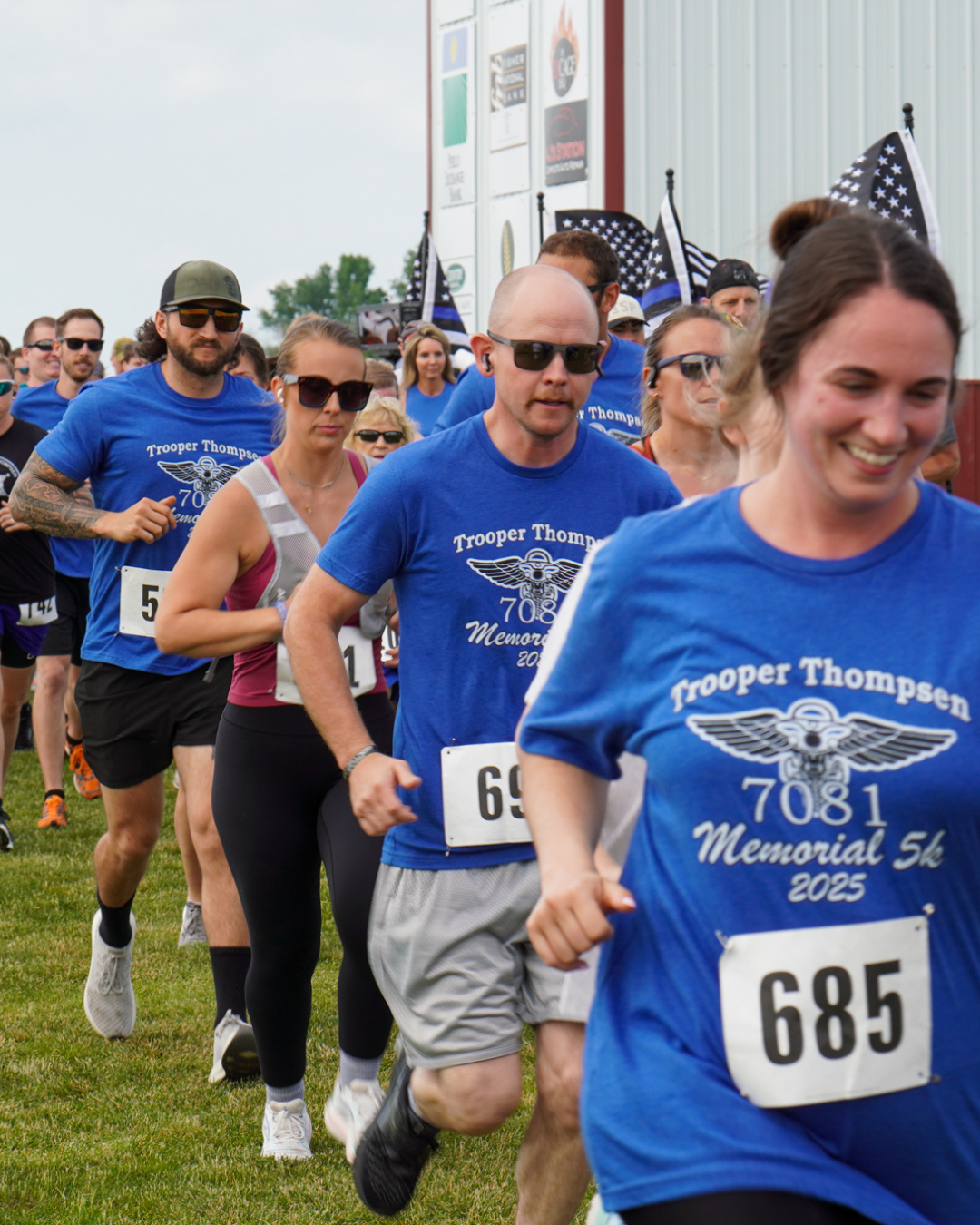 Image of 2025 Thompsen Memorial 5k racers, many of whom are running in blue race shirts and sunglasses.