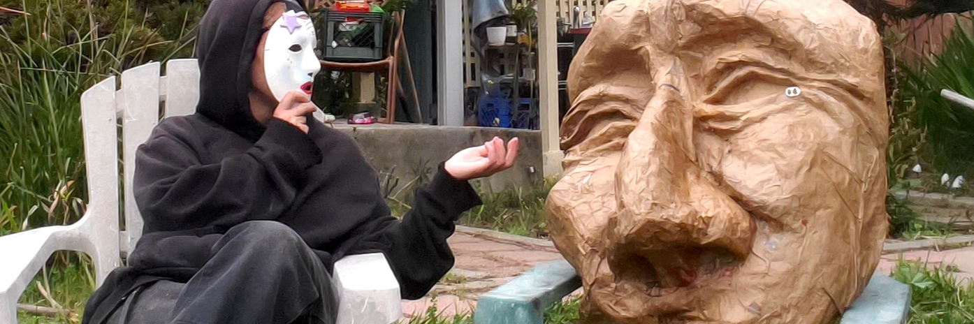 A figure in all black with a white mask sits in a lawn chair, gesturing toward a large brown paper mache mask in a lawn chair. Both sit in a grassy backyard