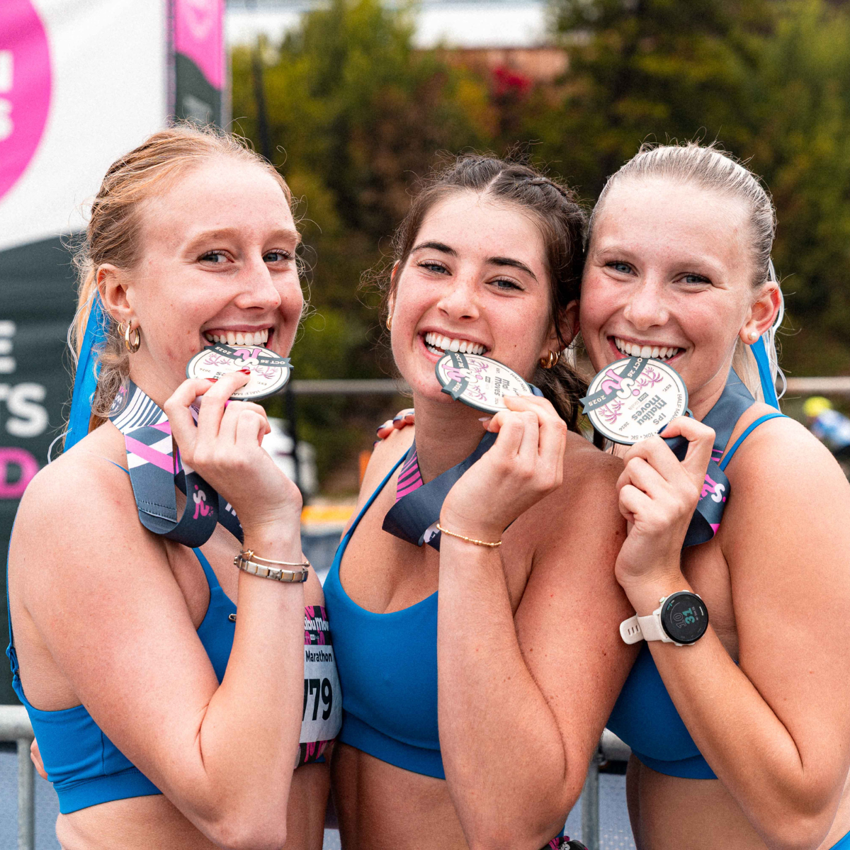 Three runners in matching blue tops pose together biting their Malibu Moves finisher medals at the post-race festival.
