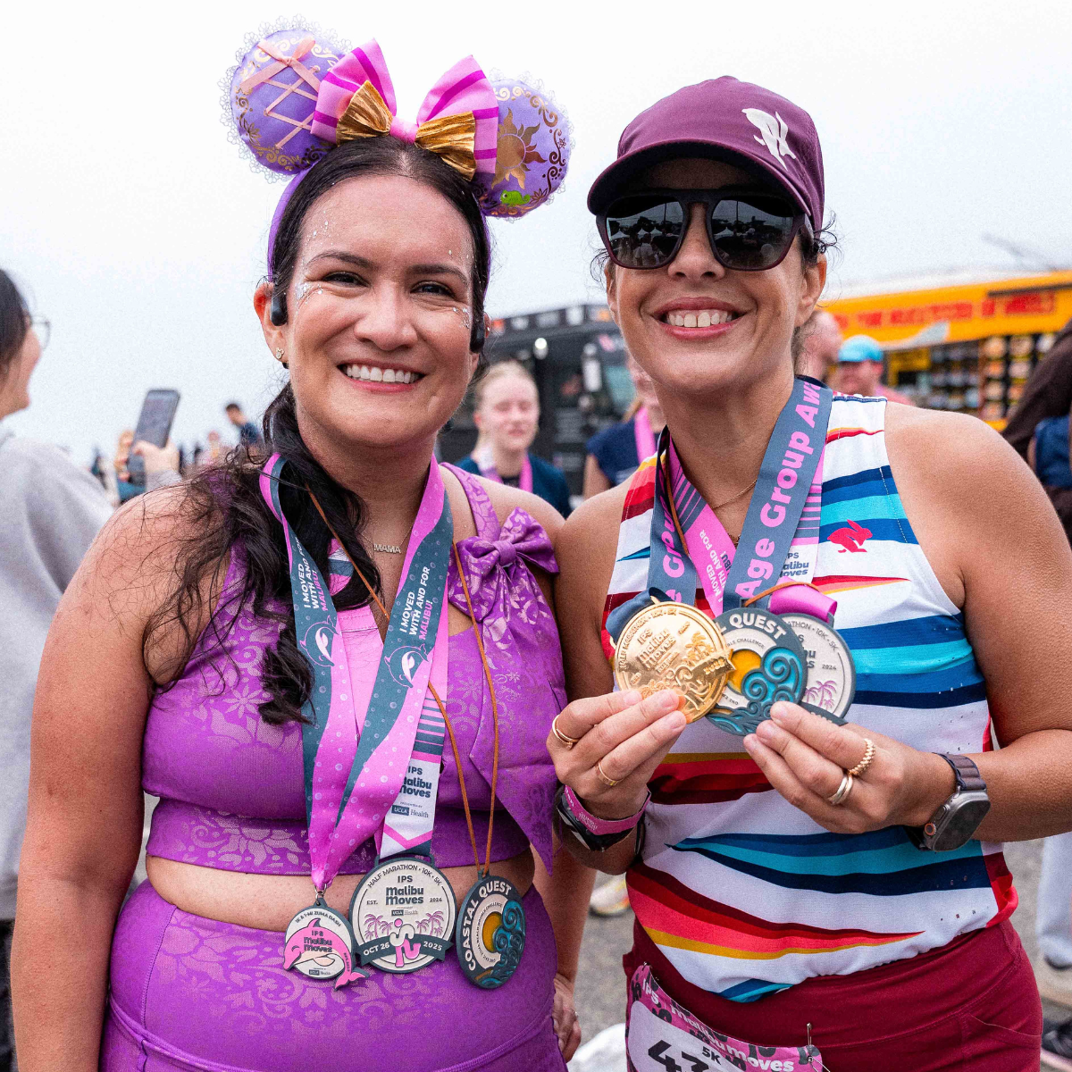 Two runners show off their collection of Malibu Moves medals, including Coastal Quest Challenge and Age Group Award hardware, at the post-race festival at Zuma Beach.