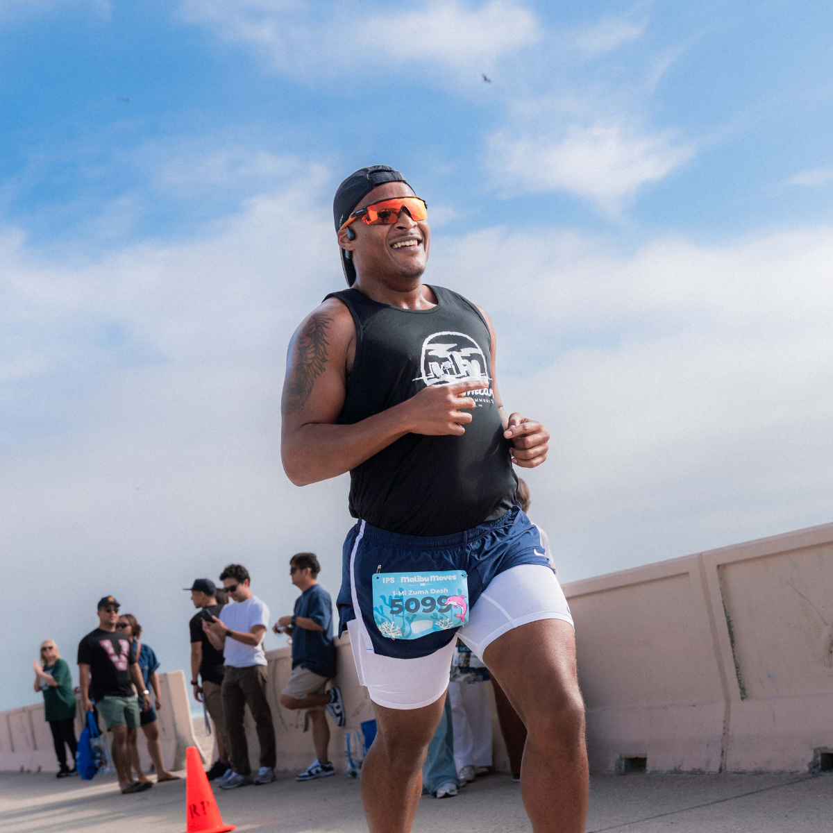 A smiling runner in orange sunglasses and a backwards cap strides through the Zuma Dash 1-Mile course with spectators and blue sky behind him.