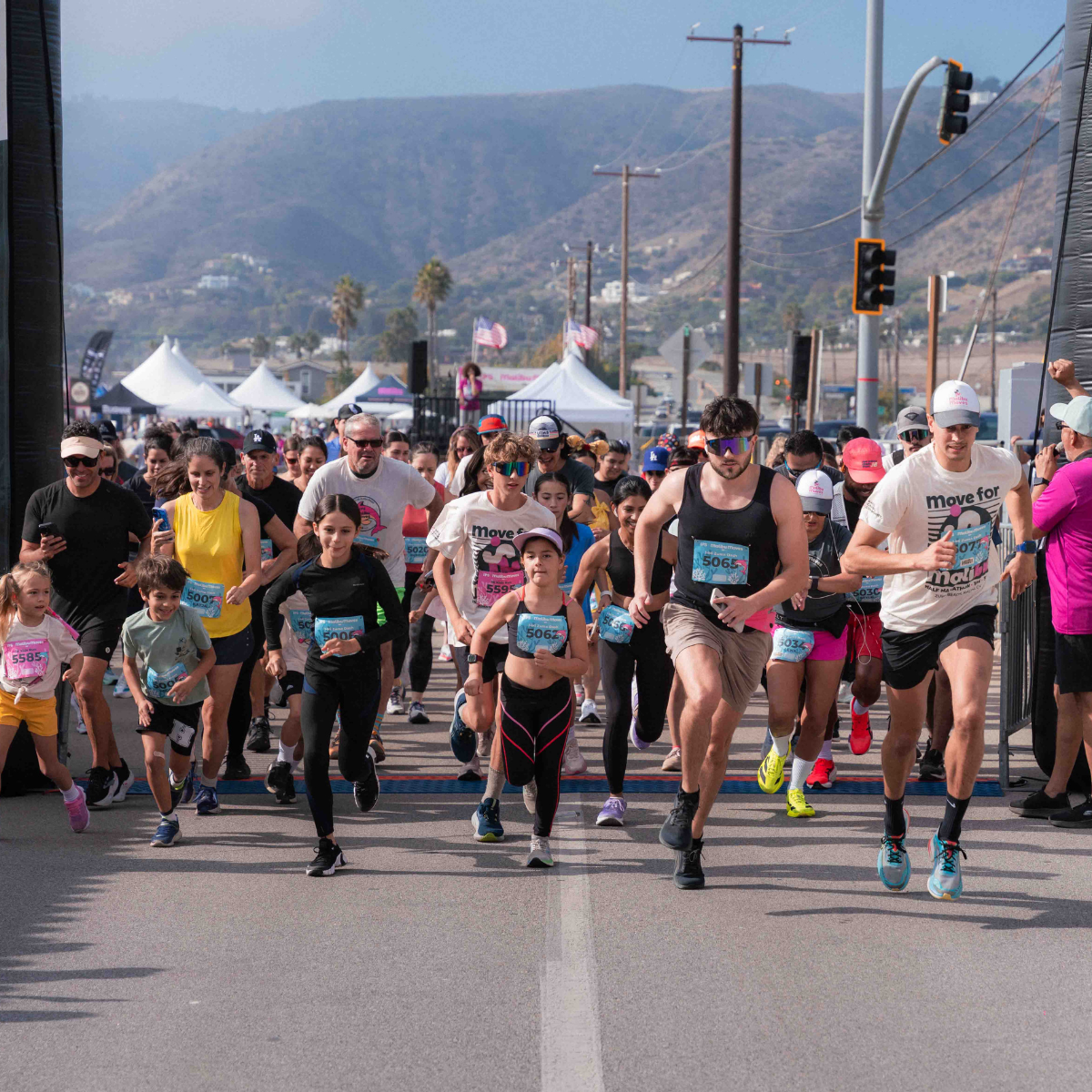 Runners of all ages charge through the Malibu Moves start line on PCH with the Malibu mountains, expo tents, and festival grounds visible in the background.