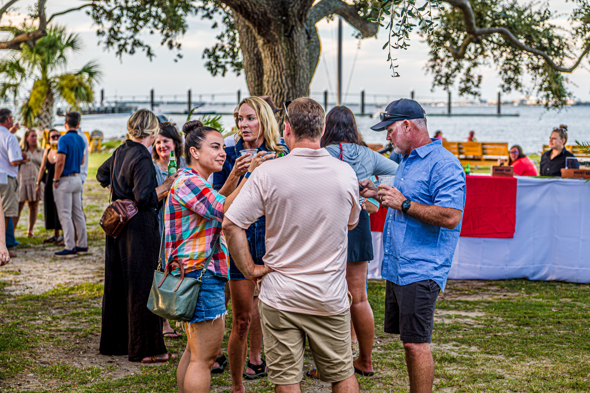 People gather under an oak tree by the water to enjoy food, drink, and live music