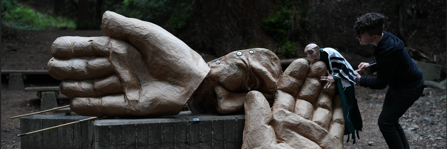 A large brown paper mache mask and two hands rest on the ground, indicating the death of the goylem. A puppeteer in all black brings the rebbe puppet towards one hand in a grieving kiss.