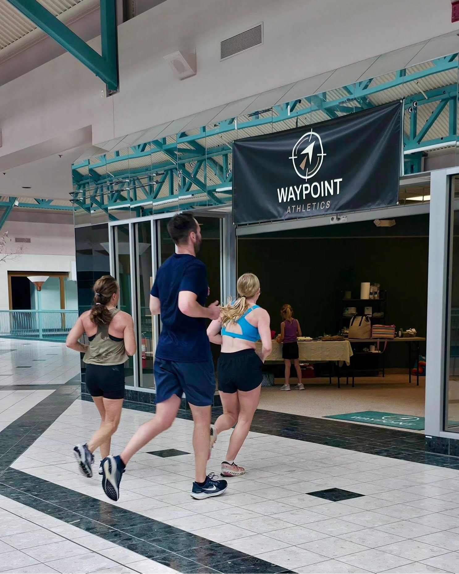 Three athletes running beneath the Waypoint Athletics banner during a gym event.