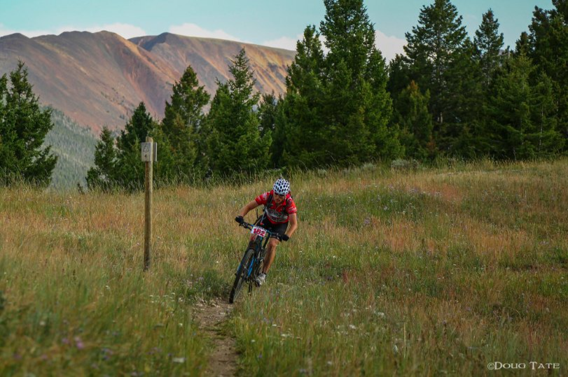 A Butte 100 racer in a red jersey rides a singletrack trail through a mountain meadow, with forested ridges behind. Photo credit: Doug Tate.